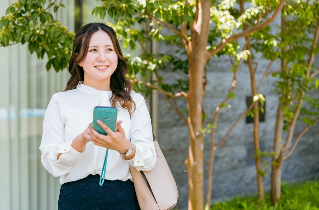 屋外でスマホを手に斜め上を見て笑顔の女性の画像